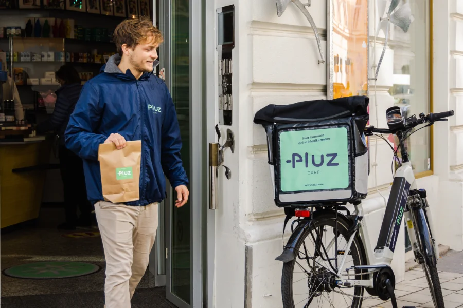 Ein Fahrer von Pluz Care geht mit einem Sackerl vom Eingang einer Apotheke zu seinem Fahrrad